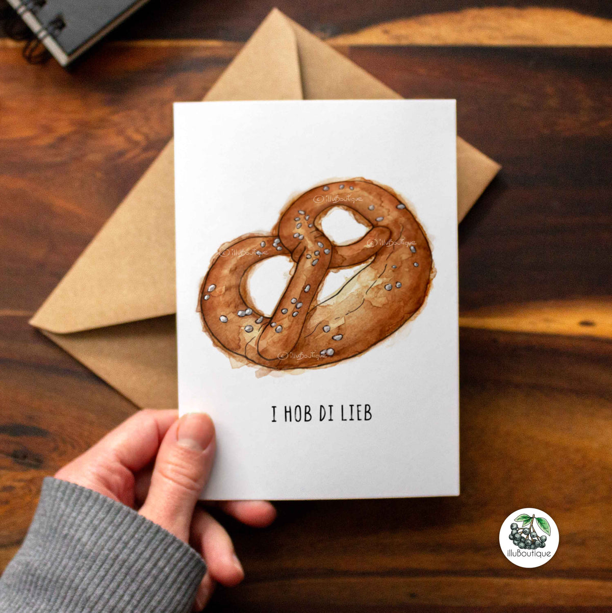 Eine weiße Karte auf einem Holztisch über einem braunen Papierumschlag mit dem Text "i hob di lieb" um im Oktoberfest Wiesn Style seine Liebe und Zuneigung zu zeigen. Darüber ein handgemaltes Aquarell Motiv einer Brezel. Über der Karte sieht man einen kleinen Teil eines Skizzenbuchs der Künstlerin und unten rechts in der Ecke das Logo von illuboutique. Kreative Idee um deine Liebe zu zeigen