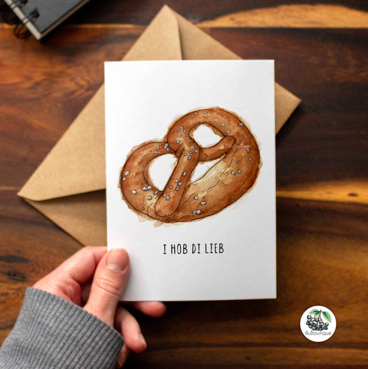 Eine weiße Karte auf einem Holztisch über einem braunen Papierumschlag mit dem Text "i hob di lieb" um im Oktoberfest Wiesn Style seine Liebe und Zuneigung zu zeigen. Darüber ein handgemaltes Aquarell Motiv einer Brezel. Über der Karte sieht man einen kleinen Teil eines Skizzenbuchs der Künstlerin und unten rechts in der Ecke das Logo von illuboutique. Kreative Idee um deine Liebe zu zeigen