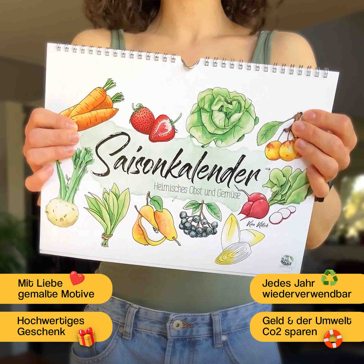 Die Künstlerin hält einen Saisonkalender in beiden Händen vor sich. Auf dem Cover sind bunte handgemalte Aquarellzeichnungen von regionalem Obst und Gemüse wie Karotten, Erdbeeren, Salat, Kohl, Kirschen und Lauch. Der Kalender trägt den Titel Saisonkalender Heimisches Obst und Gemüse. Unten links befinden sich zwei gelbe Sticker mit rotem Herz und Geschenk, die auf liebevolle Motive und hochwertige Qualität hinweisen. Geld und Co2 sparen. Jedes Jahr wiederverwendbar. Hochwertiges Geschenk