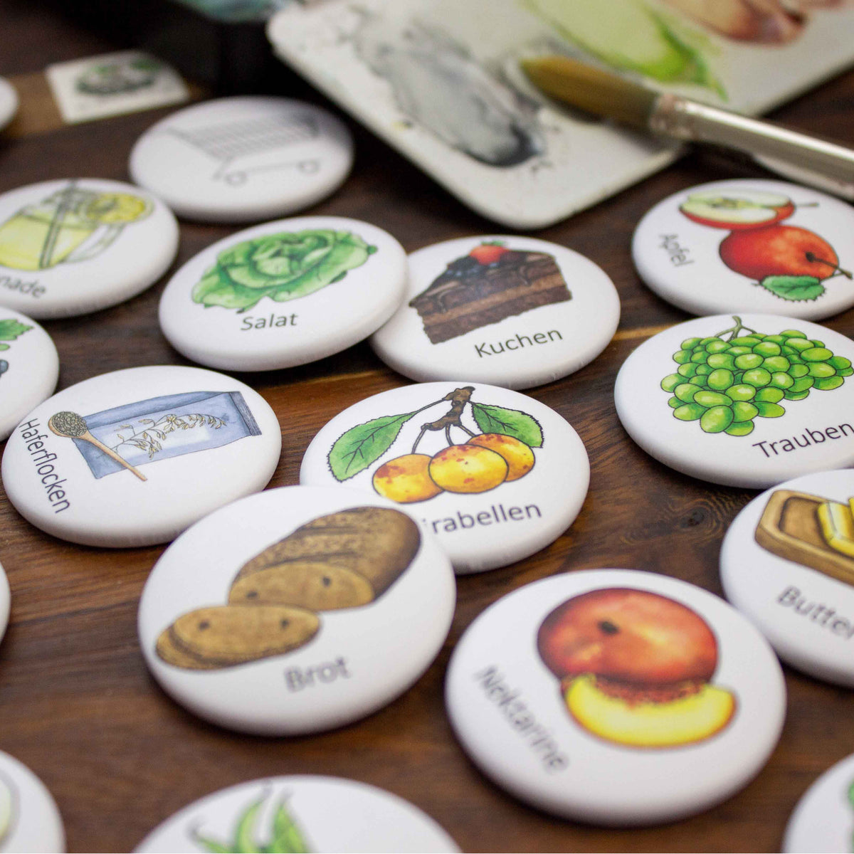 Ein Set mit weißen runden Magnet Button mit bunten Illustrationen von verschiedenen Speisen und Getränken. Die Magnete zeigen Bilder von Trauben, Kirschen, Brot, Butter, Salat, Kuchen, Limonade, Erdbeeren, Pfirsichen, Äpfeln und mehr. Die Knöpfe sind perfekt für ein Bastelprojekt oder als Lehrmittel.