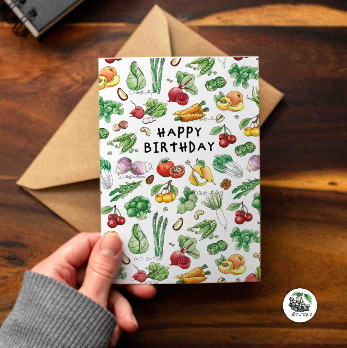 Eine weiße Karte mit bunten Motiven auf einem Holztisch über einem braunen Papierumschlag mit dem Text Happy Birthday und süßen deutschen Obst und Gemüse Bildern. Über der Karte sieht man einen kleinen Teil eines Skizzenbuchs der Künstlerin und unten rechts in der Ecke das Logo von illuboutique. Kreative Idee für runden Geburtstag Jahrgang für Mann und Frau jung und alt. Kinder freuen sich darüber genau wie Erwachsene die gerne auf Nachhaltigkeit achten oder Gemüse im Garten anbauen als Hobby haben